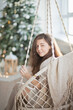© Dina - Beautiful young woman on a boho style swing near christmas tree in the living room at home, decorated for winter holiday season.