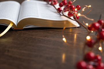 Open bible with red berries and white mini lights on a dark wood table with copy space
