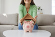 © Studio Romantic - Woman saving up money to buy her own house. Happy young girl holding roof above small pink piggy bank placed on table in living room, close up. Finance, property purchase, taking mortgage loan concept