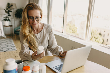 Good-looking elderly female nutrition expert sitting in living-room at table using laptop, holding bottle of vitamins writing article with advices how to select supplements for good healthy skin