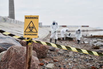 close up of warning biohazard sign at ecological disaster site with people in protective suits, copy