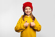 Hipster kid shows thumb up with both hands, agrees with something and says okay, like gesture, looks positively, wears yellow hoodie and red hat