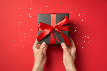 Wall Mural - First person top view photo of hands holding black giftbox with red ribbon bow and tag shiny sequins on isolated red background with text on pricetag