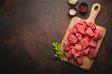 Wall Mural - Raw beef meat chopped in cubes with bunch of fresh parsley on wooden cutting board for cooking stew or other meat dish on brown dark stone concrete background top view flat lay space for text