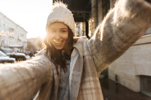 Happy Young Caucasian Girl Makes Cheerful Selfie Holding Camera With Two Hands On Background Of Sunny City. Brunette With Loose Hair In Hat Smiles With Her Teeth. Stylish Woman Having Good Time.