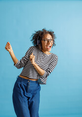 Wall Mural - Cheerful multiracial lady in headphones enjoying favorite songs and smiling while dancing in studio. Isolated on blue background