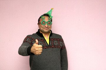 Latino adult man with Christmas hat, glasses and sweater shows his enthusiasm and happiness for the arrival of December and celebrating Christmas
