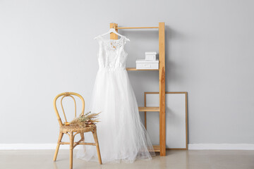 Beautiful wedding dress prepared for ceremony hanging on shelf unit and chair with bouquet of flowers near light wall