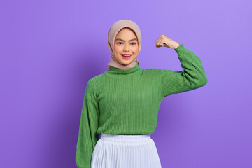 Wall Mural - Beautiful smiling Asian woman in green sweater raises arms and shows biceps, demonstrated her achievement after training in a gym isolated over purple background