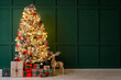 © Pixel-Shot - Beautiful Christmas tree with glowing lights, gifts and wooden reindeer near green wall