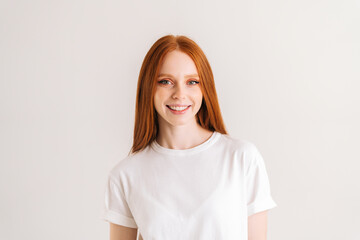 Wall Mural - Portrait of positive pretty young woman smiling and looking at camera, standing on white isolated background in studio. Happy cheerful redhead female showing sincere positive emotion.