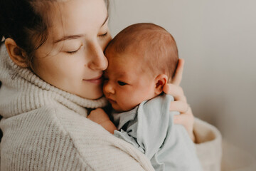 Loving mother carying of her newborn baby at home. Bright portrait of happy mum holding cute infant child on hands. Mother hugging her little 1 months old daughter.