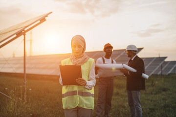 Wall Mural - Muslim woman hijab writing on clipboard while her male colleagues in helmets standing behind and examining blueprints. Multiracial people developing production of solar energy.