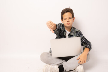 Wall Mural - Serious kid boy wearing plaid shirt sitting on floor holding laptop computer with thumb down isolated over white background.