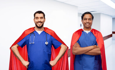 Canvas Print - healthcare, profession and medicine concept - two happy smiling doctors or male nurses in blue scrubs and red superhero capes with stethoscope over corridor at hospital on background