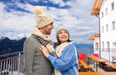 Poster - travel, tourism and winter holidays concept - happy couple in knitted hats and scarves over mountains and ski resort background