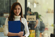 © saltdium - Photo of cheerful woman owner of coffee shop in apron standing at the entrance over the open signboard as background.