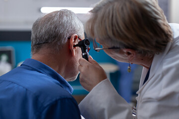 Wall Mural - Woman doctor holding otoscope to do ear consultation for old man at medical appointment. Otologist using otology instrument to do examination and give medical advice to ill patient.