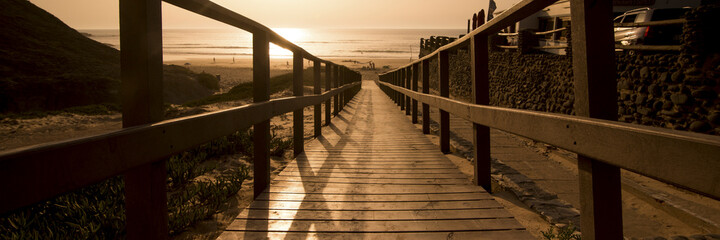 Wall Mural - Horizontal banner header landscape picture with empty wooden pier and beautiful beach at sunset time with golden ocean water in background. Concept of travel and summer holiday vacation