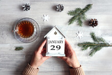 Calendar For January 29: Hands Hold A Decorative House With The Name Of The Month And The Number 29, A Cup With Hot Tea, Snowflakes, Fir Branches On A Gray Background, Top View.