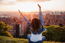 Woman With Arms Raised Looking At City While Sitting In Park During Sunset
