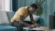 African American guy in glasses calculating utility bills and taxes and getting upset while sitting on sofa at home