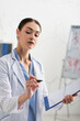 © LIGHTFIELD STUDIOS - brunette chemist holding clipboard while writing formulas on glass board.
