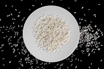 Wall Mural - Rice in a white plate and grains spilled on a black background.