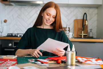 Medium shot portrait of happy young woman cutting out craft paper for envelopes with scissors making Christmas advent calendar on winter holidays eve, selective focus, blurred background.