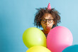 Young lady in birthday cone hat pouting lips and making funny face while holding colorful balloons. Isolated on blue background