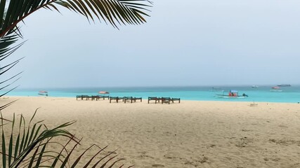 Wall Mural - Azure water with boats and the beach of the Indian ocean in bad rainy weather. Seaview through a palm leaf