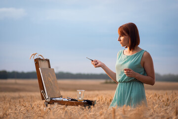 Wall Mural - Female painter during work outdoors. Red-headed girl artist making last brushstrokes on her drawing, pleased on the result of her work.