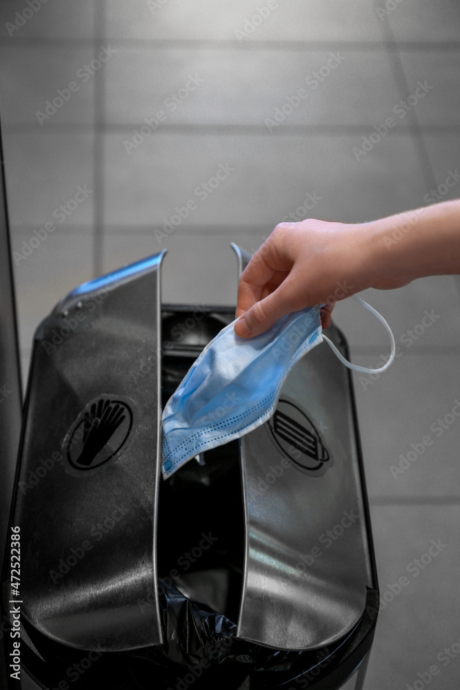 Woman throwing used medical mask into garbage bin indoors