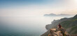 © YURII Seleznov - A young Asian woman with a backpack hiking in the summer sits on top of a montage and looks out over a beautiful sea bay landscape. Mountain and coastal travel, freedom and an active lifestyle