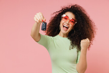 Poster - Exultant happy excited young curly latin woman 20s wear mint t-shirt sunglasses hold vehicle key doing winner gesture isolated on plain pastel light pink background studio portrait. Car sales concept