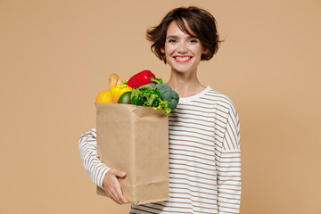 Wall Mural - Young smiling european cheerful happy fun vegetarian woman 20s in casual clothes hold paper bag with vegetables after shopping look camera isolated on plain pastel beige background studio portrait