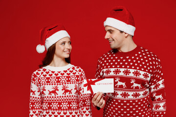 Wall Mural - Young fun couple friends two man woman in sweater hat hold gift store certificate coupon voucher card isolated on plain red background. Happy New Year 2022 celebration merry ho x-mas holiday concept.