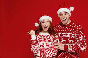 Wall Mural - Young couple friends two man woman 20s in sweater hat point finger aside on copy space isolated on plain red background studio portrait. Happy New Year 2022 celebration merry ho x-mas holiday concept.