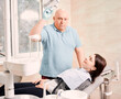 © anatoliy_gleb - Male stomatologist looking at camera while standing beside patient. Woman lying in dental chair while having appointment with dentist and waiting for procedure. Concept of dentistry and stomatology.