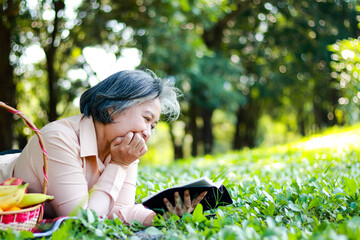 Wall Mural - Asian elderly woman lying in the park reading a book on the grass She is enjoying her vacation. The concept of living in retirement to be happy. Copy space