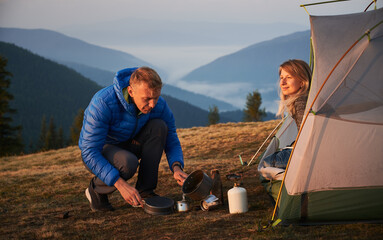Wall Mural - Camping in the morning. Hiker man sitting squatting and pouring water to make coffee near his wife, who admiring the surrounding beauty at dawn from their tent against misty mountains.