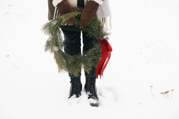 Winter Holidays in countryside. Stylish woman with christmas wreath in winter park. Young female in cozy mittens and coat holding xmas wreath with red bow in snowy winter field, cropped view