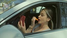 Bored Young Woman Stuck In Traffic.Young Woman Dissatisfied With Traffic In The City, Using Her Mobile Phone As A Mirror, Applying Lipstick To Her Lips. Makeup Concept In Traffic.