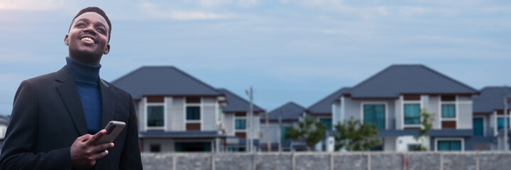 African smiling businessman stands in front of a housing estate while using a smartphone.Concept of Realty business