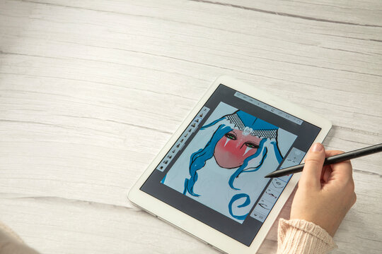 Young creative designer holding stylus pen drawing on screen of digital tablet on wooden desk with copy space, modern digital art top view