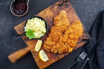 Traditional deep-fried schnitzel with potato salad and cranberries served as top view on an old rustic wooden board