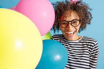 Wall Mural - Close up of cheerful multiracial lady looking at camera and smiling while holding colorful balloons. Isolated on blue background