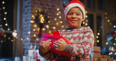 Little multiracial boy wearing Santa hat holding gift box with present while spending time at home with New Year decorations. Christmas spirit concept