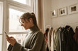 Profile photo of smiling girl with phone in hands stands at window in dressing room of house. Lady with gathered hair wears glasses, headphones and jacket. Telephone communication technology concept