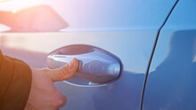 Driver in warm black jacket uses keyless entry access by putting finger on handle to open or close blue foreign car with fingerprint closeup, sunlight